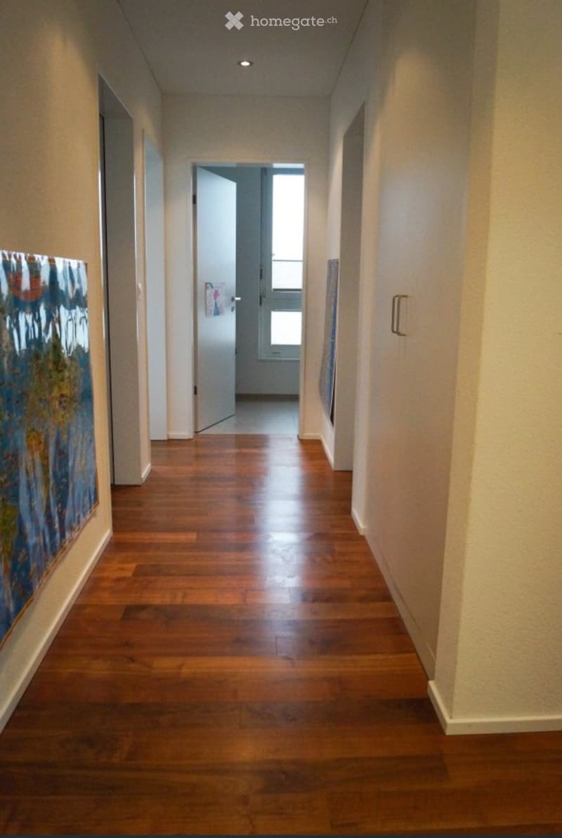 A long, well-lit hallway with wooden flooring, white walls, and a door at the end leading to another room.