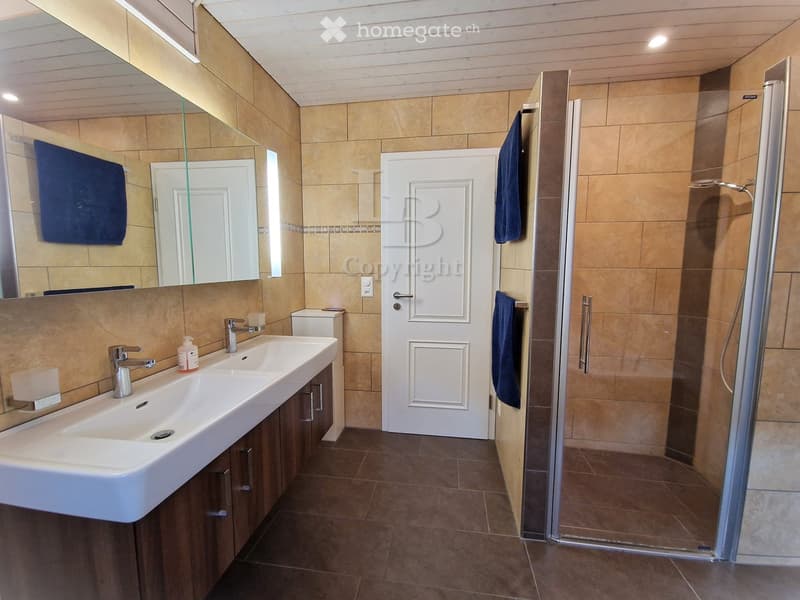 Modern bathroom with beige tiles, a double sink, a large mirror, a glass-enclosed shower, and blue towels.