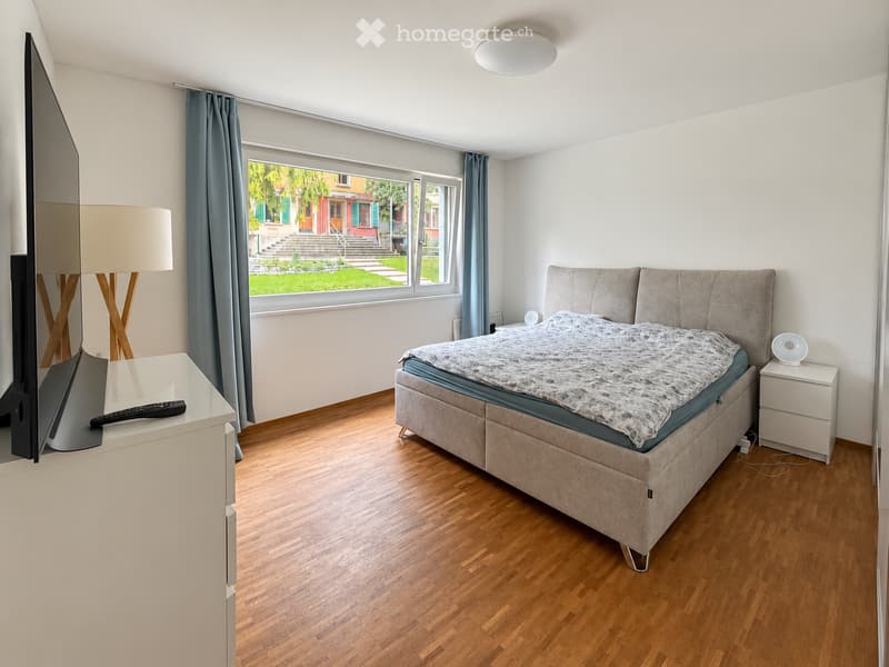 Modern bedroom with wooden flooring, large window with blue curtains, bed with headboard, nightstand, lamp, and TV on a stand.