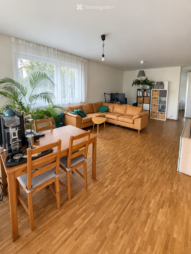 A modern living room with wooden flooring, a large window with white curtains, a brown leather sofa, a wooden dining table with chairs, a coffee machine, a screen, and a lamp.