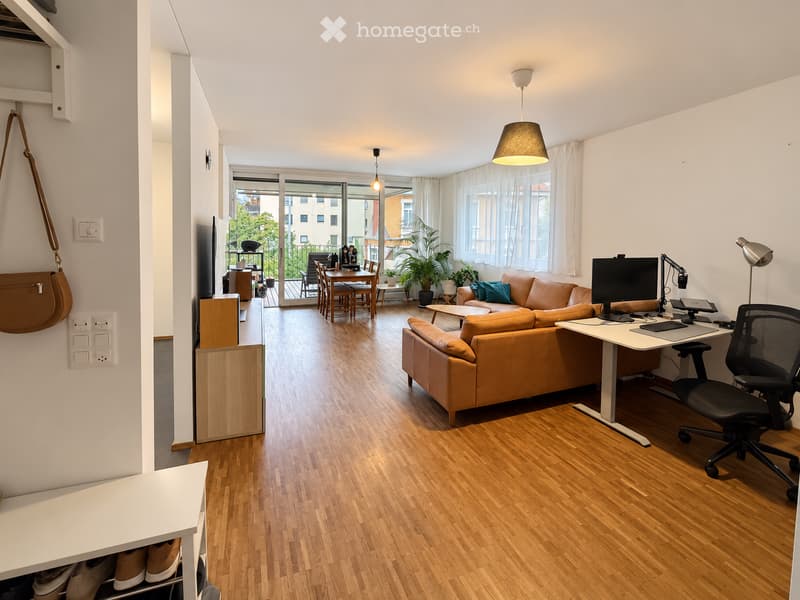 Open-plan living and dining area with wooden flooring, a brown leather sofa, a dining table with chairs, a desk with a computer, and a balcony with outdoor seating.