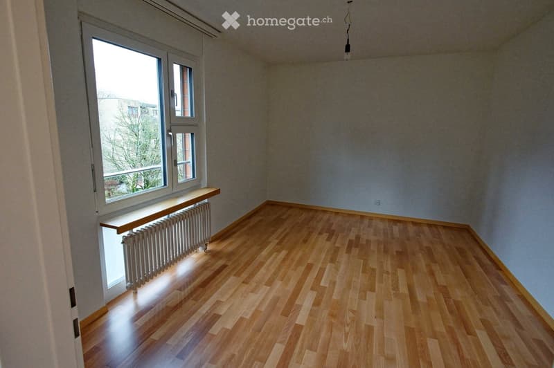 Empty room with wooden flooring, light-colored walls, large window with a radiator below, and a small shelf on the window sill.