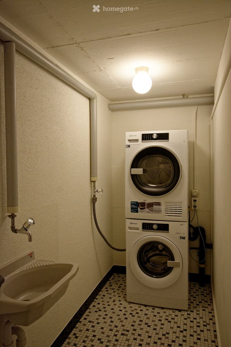 A small bathroom with a washing machine and a sink, featuring tiled flooring and white walls.