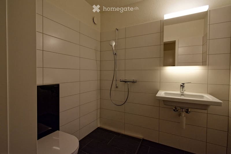 Modern bathroom with white tiled walls, a toilet, a sink with a modern faucet, a showerhead, and a built-in mirror with integrated lighting.