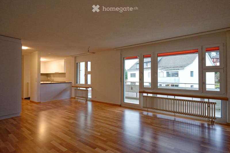 Empty room with wooden flooring, large windows, radiators, and a small kitchen area visible in the background.
