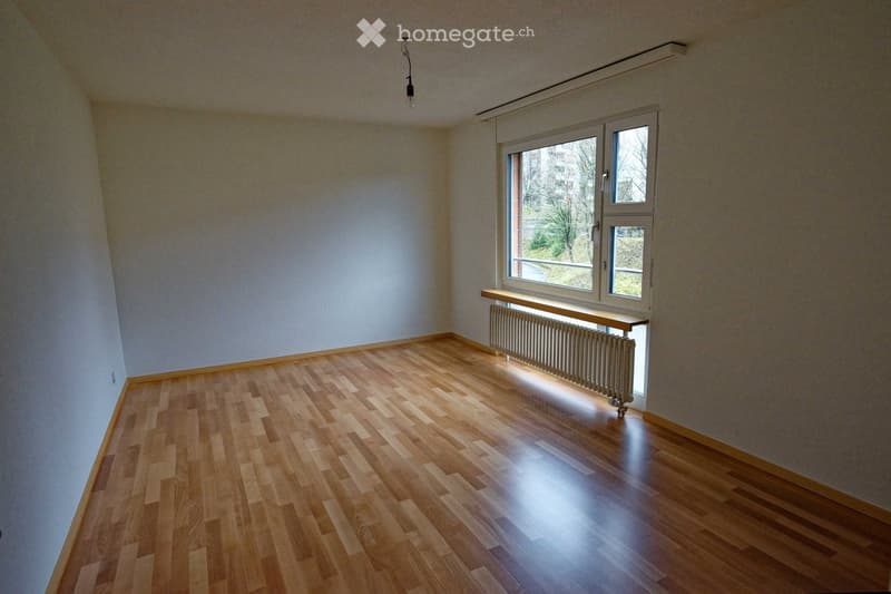 Empty room with wooden flooring, light-colored walls, and a window with a radiator below it. No furniture or personal items are present.