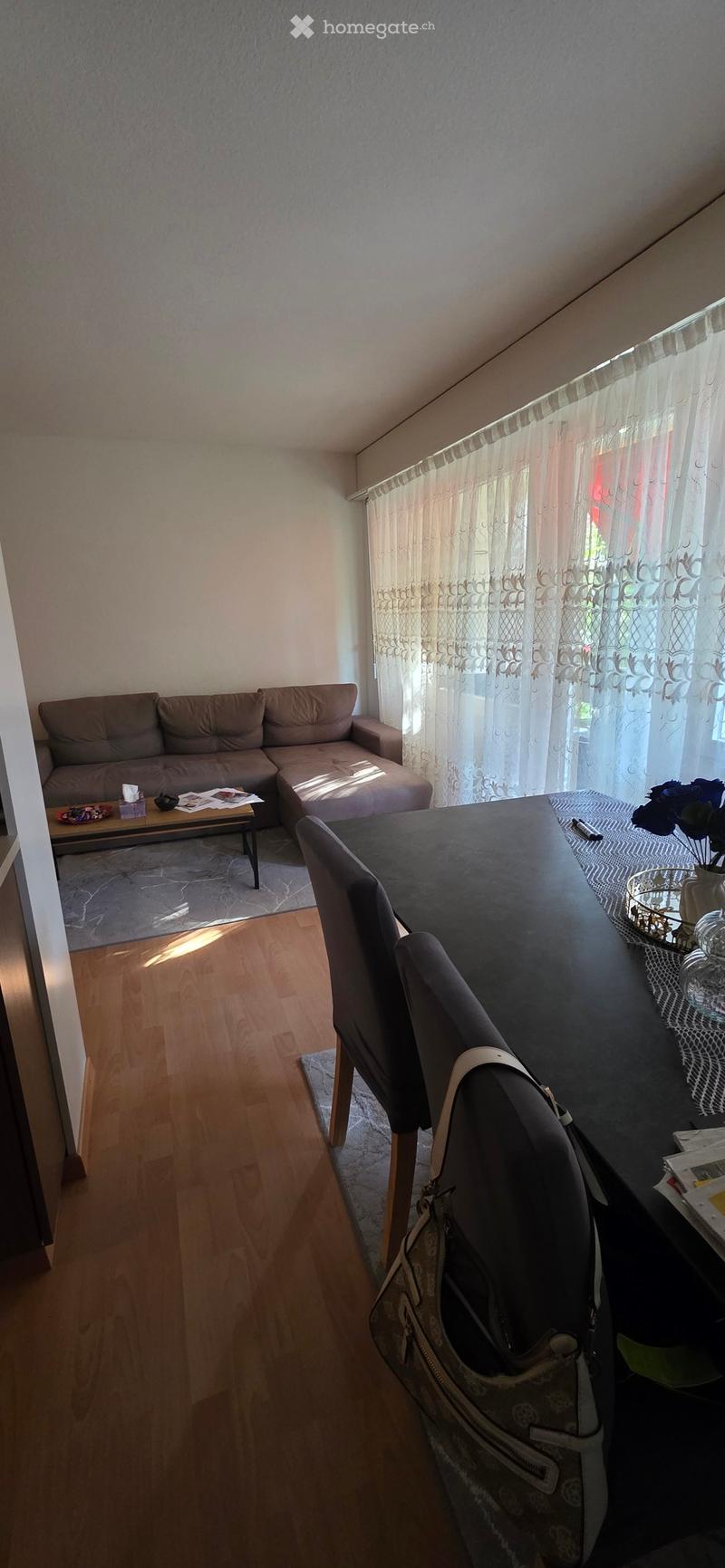 A modern living room with a sectional sofa, coffee table, dining table, chairs, and a vase on the table. Natural light filters through sheer curtains.
