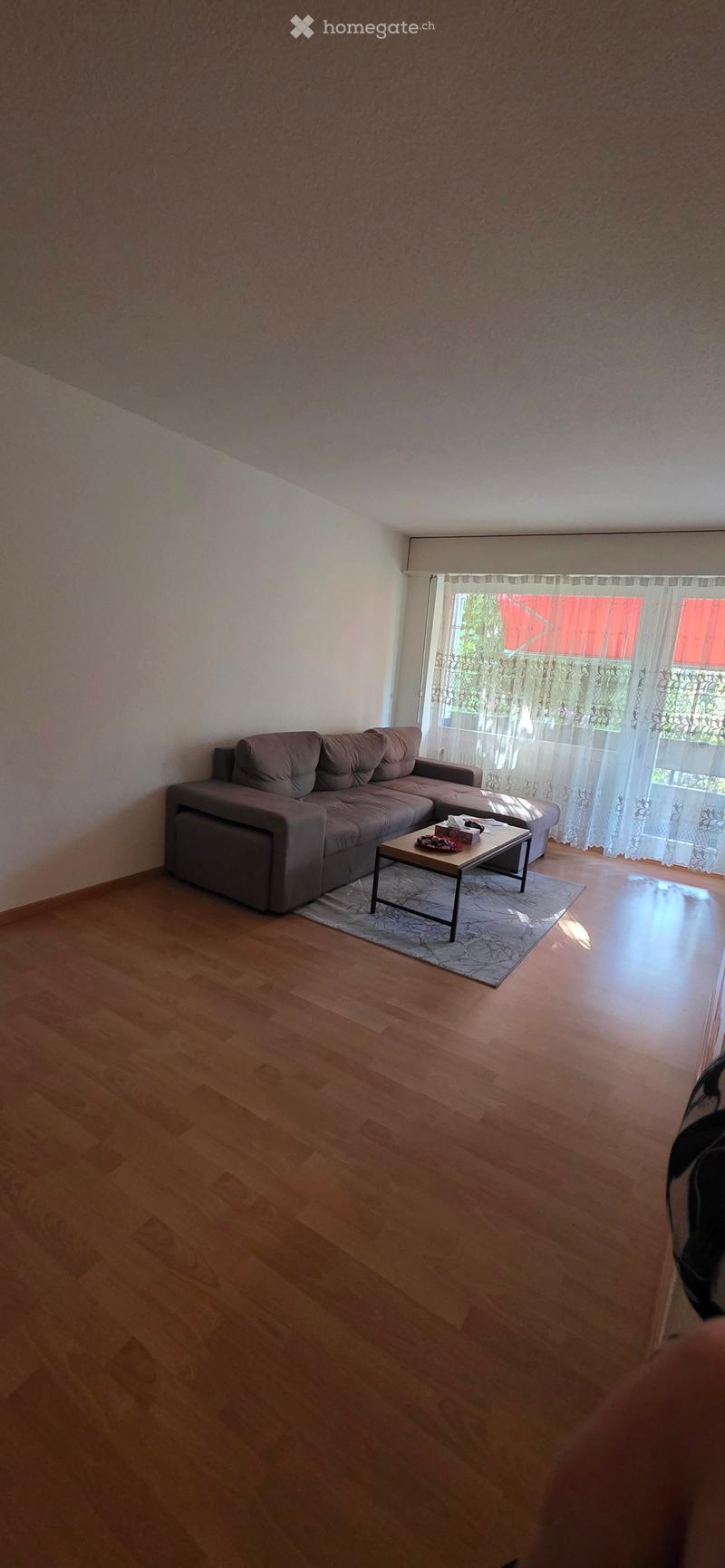 Living room with wooden flooring, grey sectional sofa, small coffee table, and large window with sheer curtains.