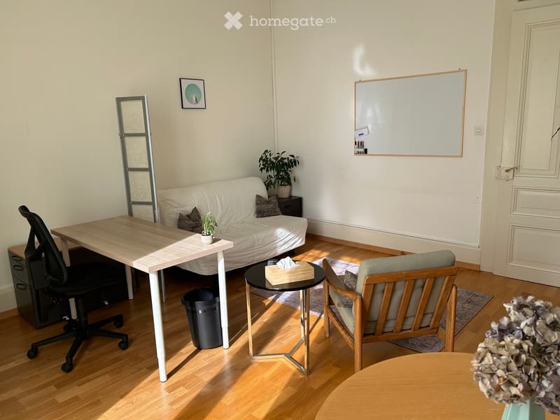 A modern, well-lit living space with a white sofa, wooden table, office chair, and small side table with a plant. The room features a whiteboard, picture frame, and a small plant on the table.