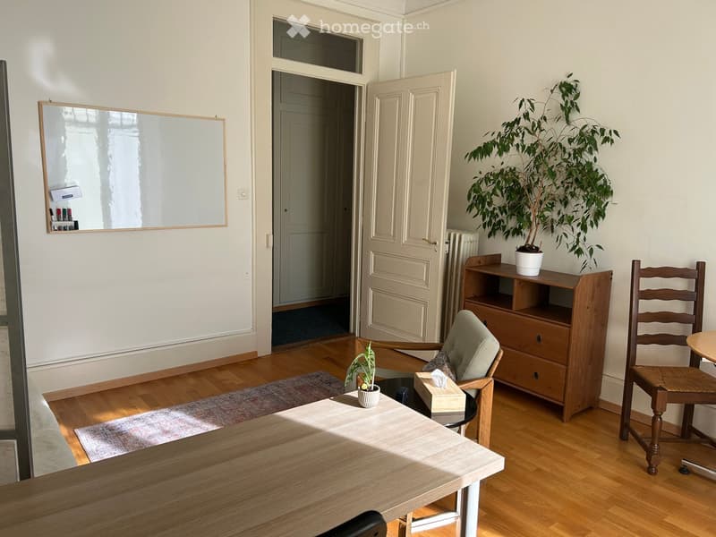 A furnished room with a wooden table, chairs, a cabinet, and plants. The room features wooden flooring, a large mirror on the wall, and an open doorway leading to another space.