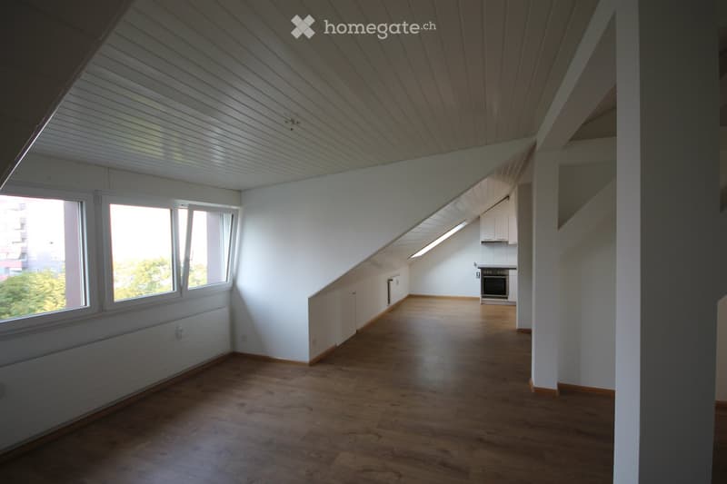 Bright, empty room with wooden flooring, white walls, and large windows offering a view of greenery. The room features a slanted ceiling and a small kitchen area visible in the background.