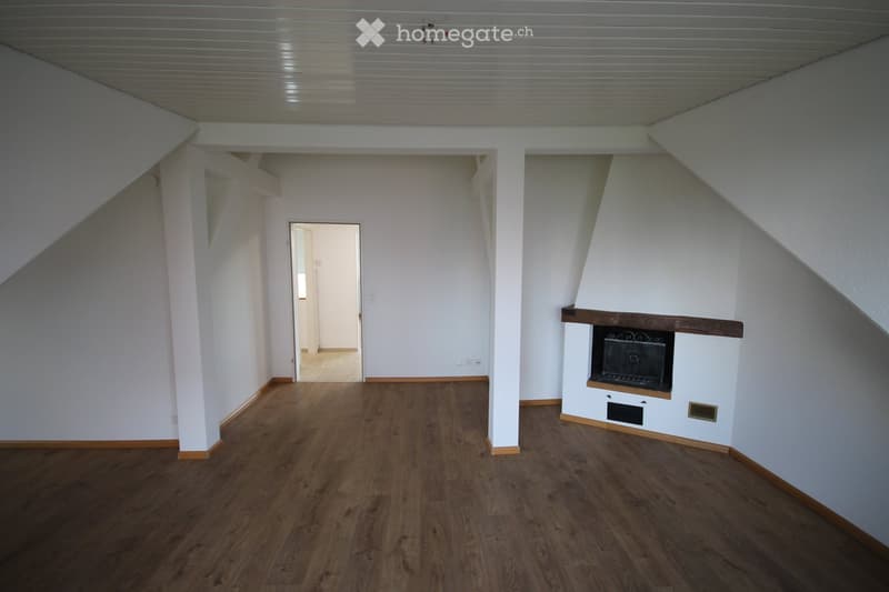 Empty room with wooden flooring, white walls, and a fireplace in the corner. The room has a slanted ceiling and a doorway leading to another space.