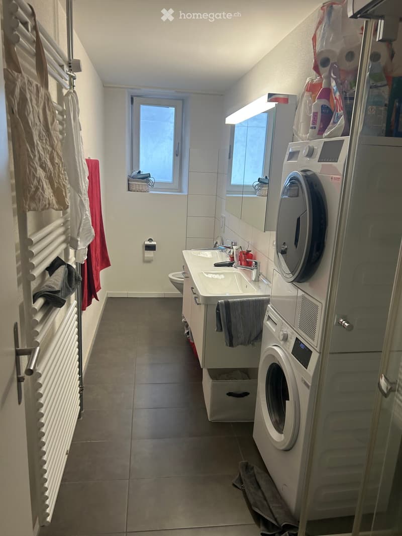 Modern bathroom with a double sink, toilet, washing machine, radiator, and a window with a view.