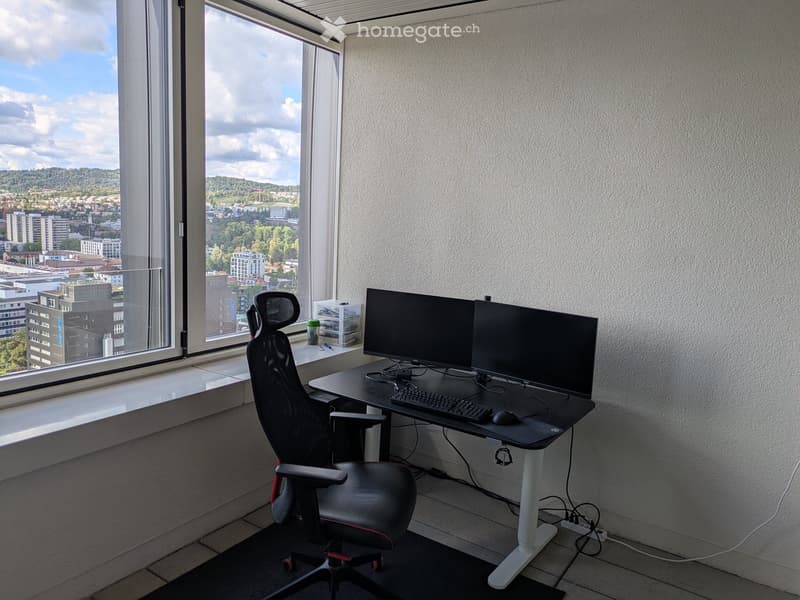 Modern office space with large windows offering a city view, featuring a black ergonomic chair, a standing desk with dual monitors, keyboard, and mouse.