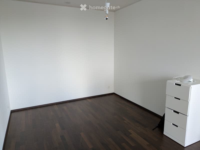 Empty room with white walls and dark wooden flooring, featuring a single white storage unit with drawers and a small electronic device on top.