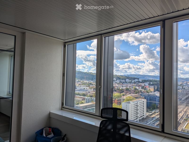 Office space with large windows offering a cityscape view, featuring a single chair and a small table with items.