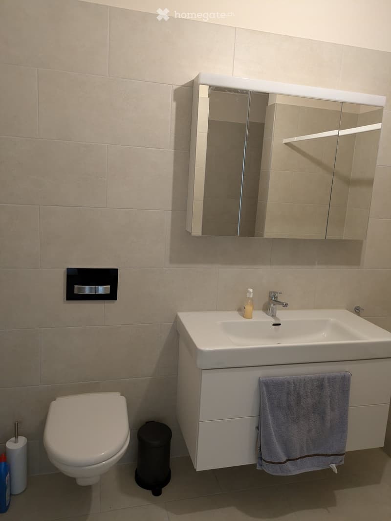 Modern bathroom with beige tiles, a white sink with a mirror cabinet, a toilet, and a towel hanging on the cabinet.