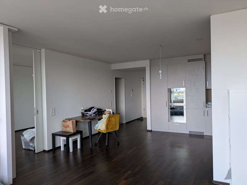 A modern, minimalist room with a dark wooden floor, white walls, and built-in cabinets. A small table with a chair and a stool is placed near the wall, with a shopping bag and some items on the table. An oven and refrigerator are visible in the background