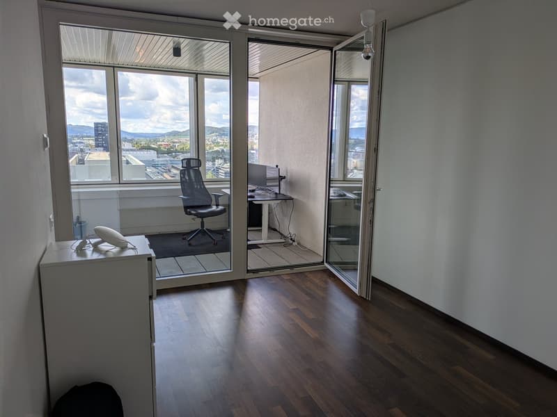 Office space with wooden flooring, large windows offering a city view, a desk with a computer, a chair, and a small cabinet with a phone.