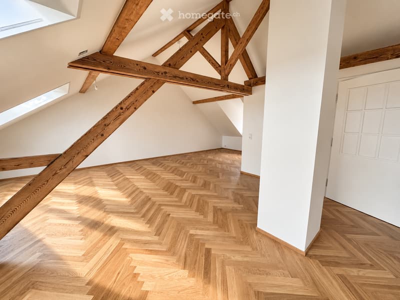 Bright, empty attic room with exposed wooden beams, herringbone wooden flooring, and skylights allowing natural light to fill the space.