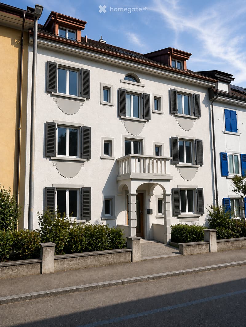 A two-story white house with multiple windows, shutters, and a small balcony on the first floor. The house features decorative moldings and is surrounded by low hedges and a paved walkway.