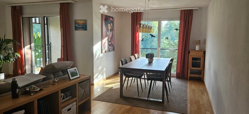 Bright living and dining area with wooden flooring, large windows offering a scenic view, a sofa, dining table with chairs, a side cabinet, and decorative items.
