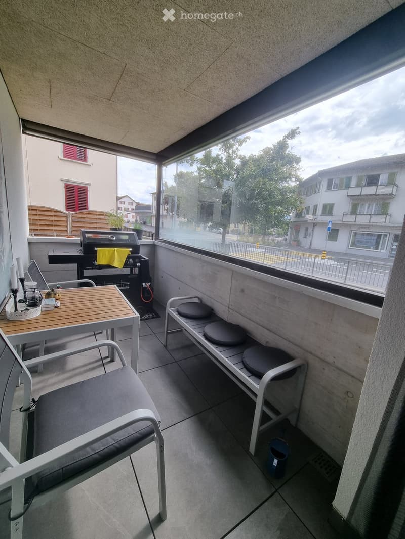 Balcony with tiled floor, wooden table, white chairs, bench with stools, grill, and small candle holder.