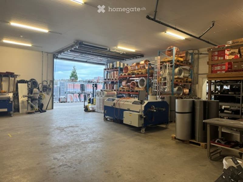 Large industrial garage with shelving units, machinery, and storage bins. The space is well-lit with fluorescent lighting and has a concrete floor.