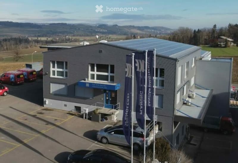 Modern industrial-style building with solar panels on the roof, multiple windows, and a blue entrance sign. Several vehicles are parked in the lot.