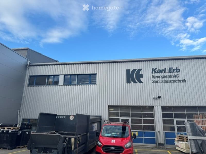 Industrial building with a modern facade, large windows, and signage for "Karl Erb Spengler AG San. Haustechnik". A red van is parked in front, and there are containers and pallets nearby.
