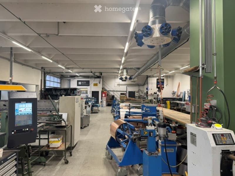Industrial workshop with various machinery, workstations, and tools. The space features a high ceiling with exposed ductwork and fluorescent lighting. Machines and equipment are arranged in rows, with a computer monitor visible on a cart.