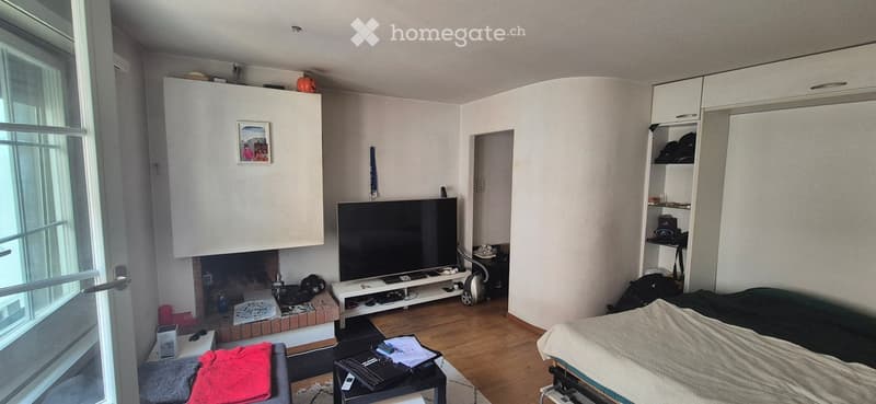 Modern living room with a fireplace, TV stand, sofa, coffee table, bed, and cabinet. The room features a large window and a wooden floor.