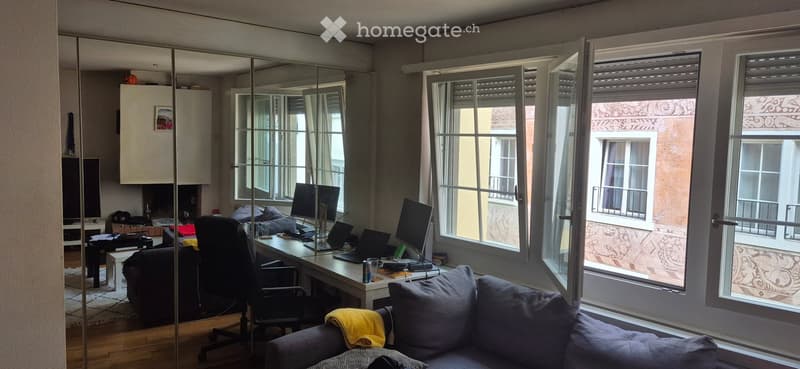 A modern, well-lit room with large windows, a sofa, a desk with a laptop and screen, a chair, and a mirror. The room features a mix of contemporary and functional furniture.