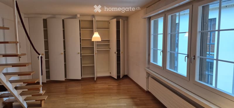 Empty room with wooden flooring, large windows, built-in white cabinets, and a modern staircase with a rope handrail.