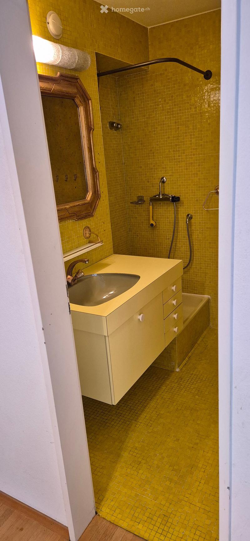 Bright yellow tiled bathroom with a sink, mirror, and bathtub. The walls and floor are covered in small yellow tiles, and the sink has a modern faucet.