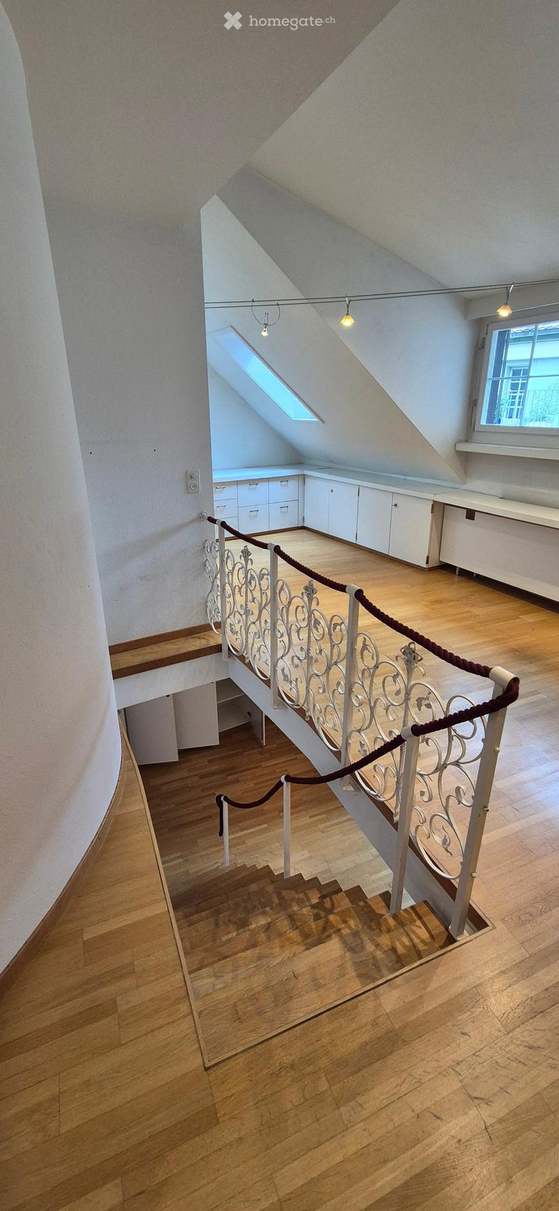Wooden staircase with white ornate railing and rope handrail, leading to an attic space with white cabinets and a skylight window.