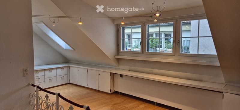 Bright, empty attic room with wooden flooring, white built-in cabinets, large windows, and a skylight. The room features exposed wooden beams and modern lighting fixtures.