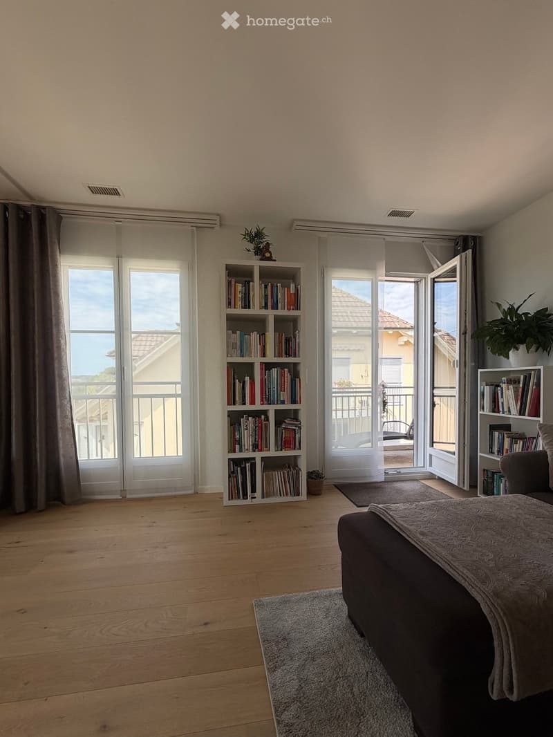 Bright living room with wooden flooring, large windows, and a balcony. Features a sofa, bookshelf, and plants.