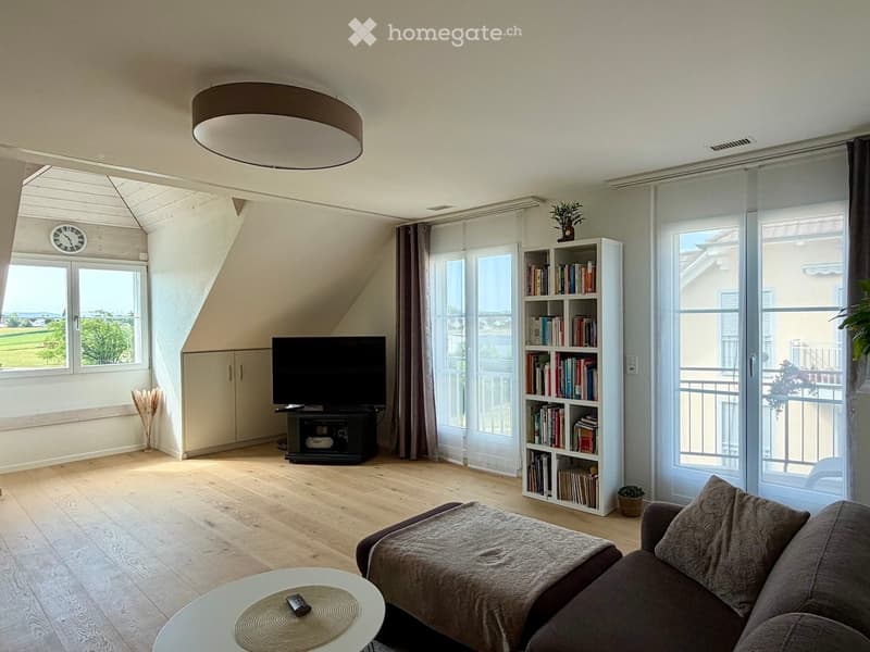 Bright living room with wooden flooring, large windows offering a scenic view, a sofa, coffee table, television, bookshelf, and a clock on the wall.
