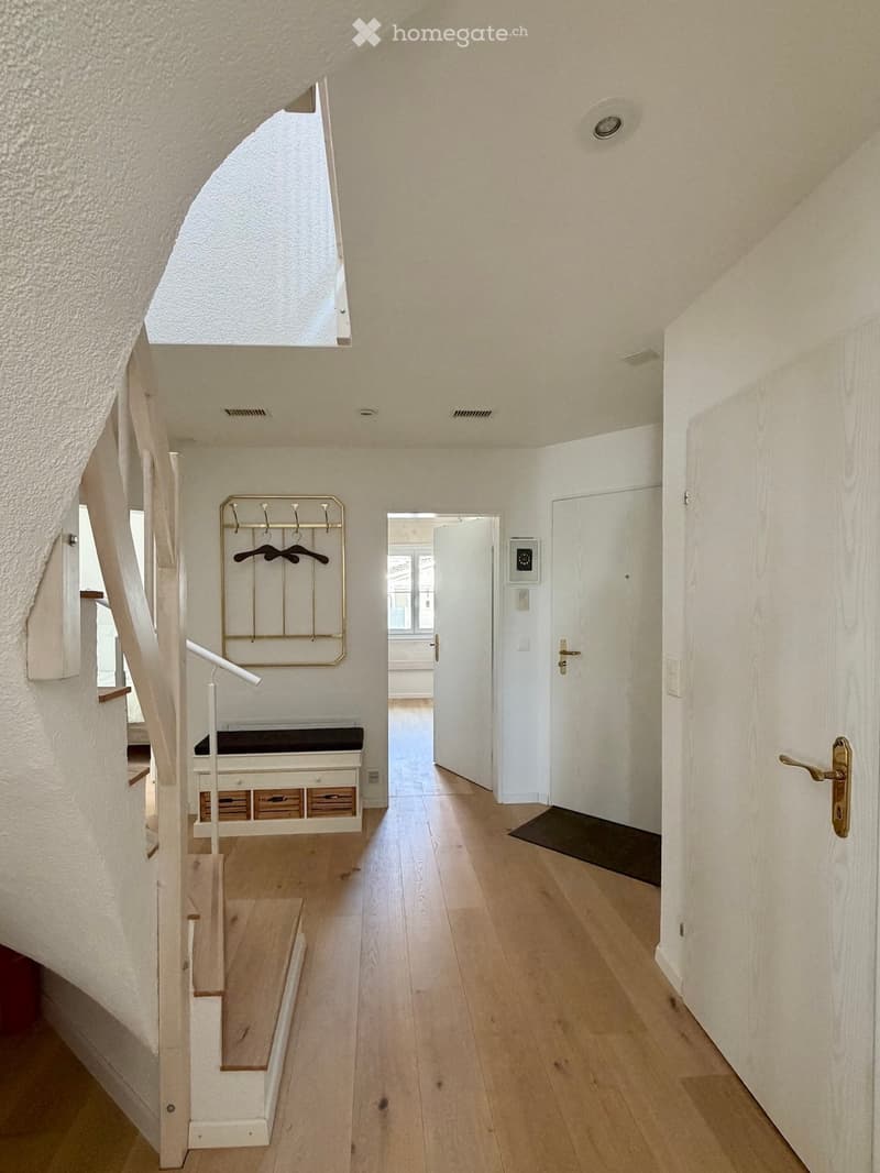 Bright hallway with wooden flooring, white walls, and a staircase leading upwards. Features include a coat rack, a bench with drawers, and a small mat near a door.