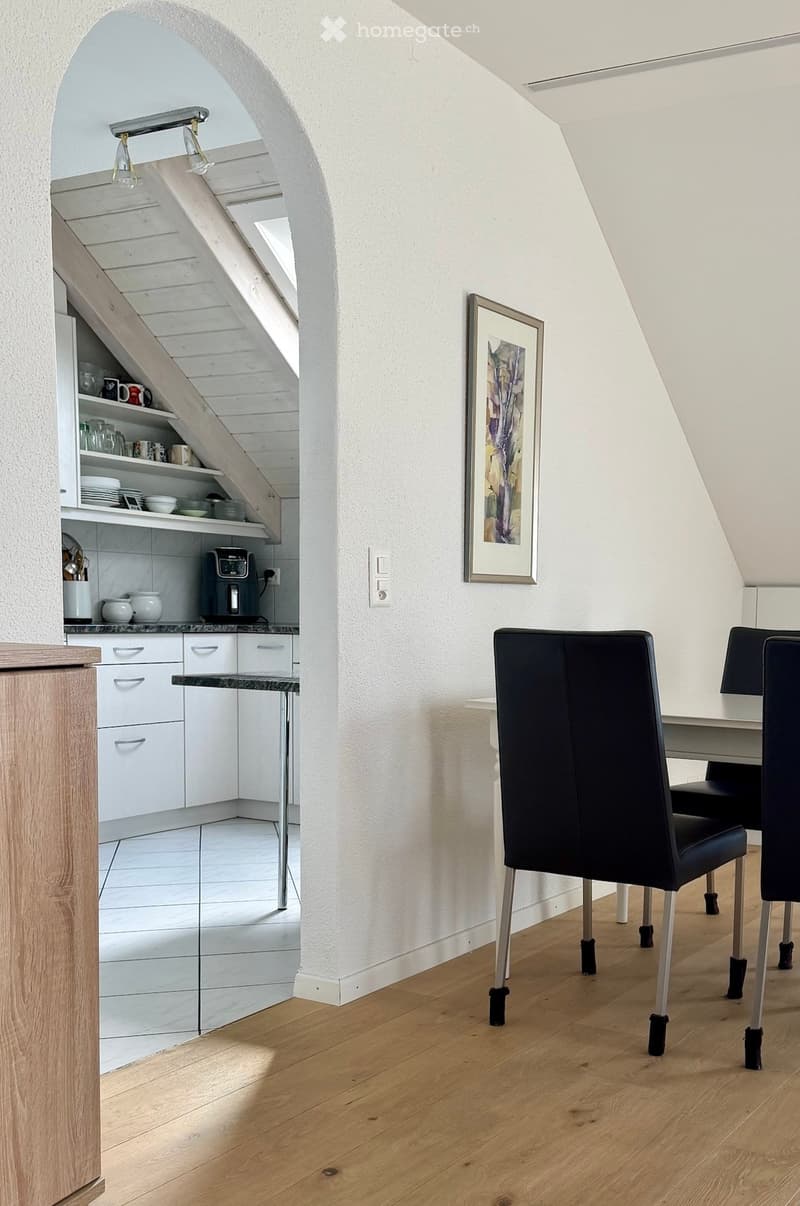 A modern, open-plan living and dining area with a wooden floor, white walls, and a staircase leading to an upper level. The space includes a dining table with black chairs, a small kitchen area with white cabinets, a refrigerator, and various dishes and c