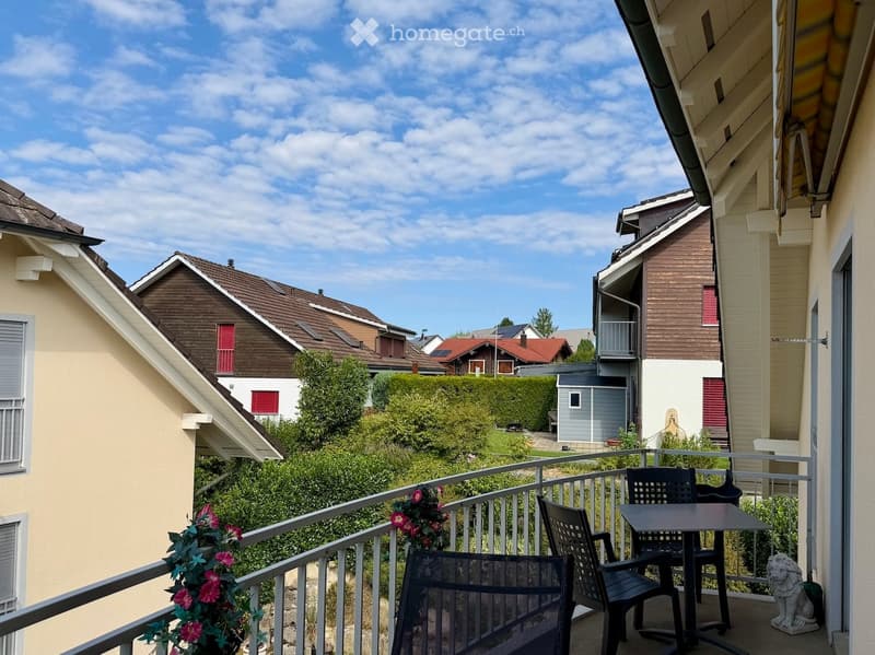 Balcony with a metal railing, a small table, two chairs, and a flower pot with pink flowers. The view shows a residential neighborhood with houses, greenery, and a clear blue sky.