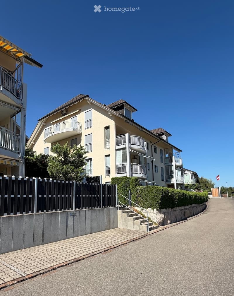 A two-story residential house with a light yellow facade, multiple balconies, and a well-maintained garden area with a low hedge and a black fence.
