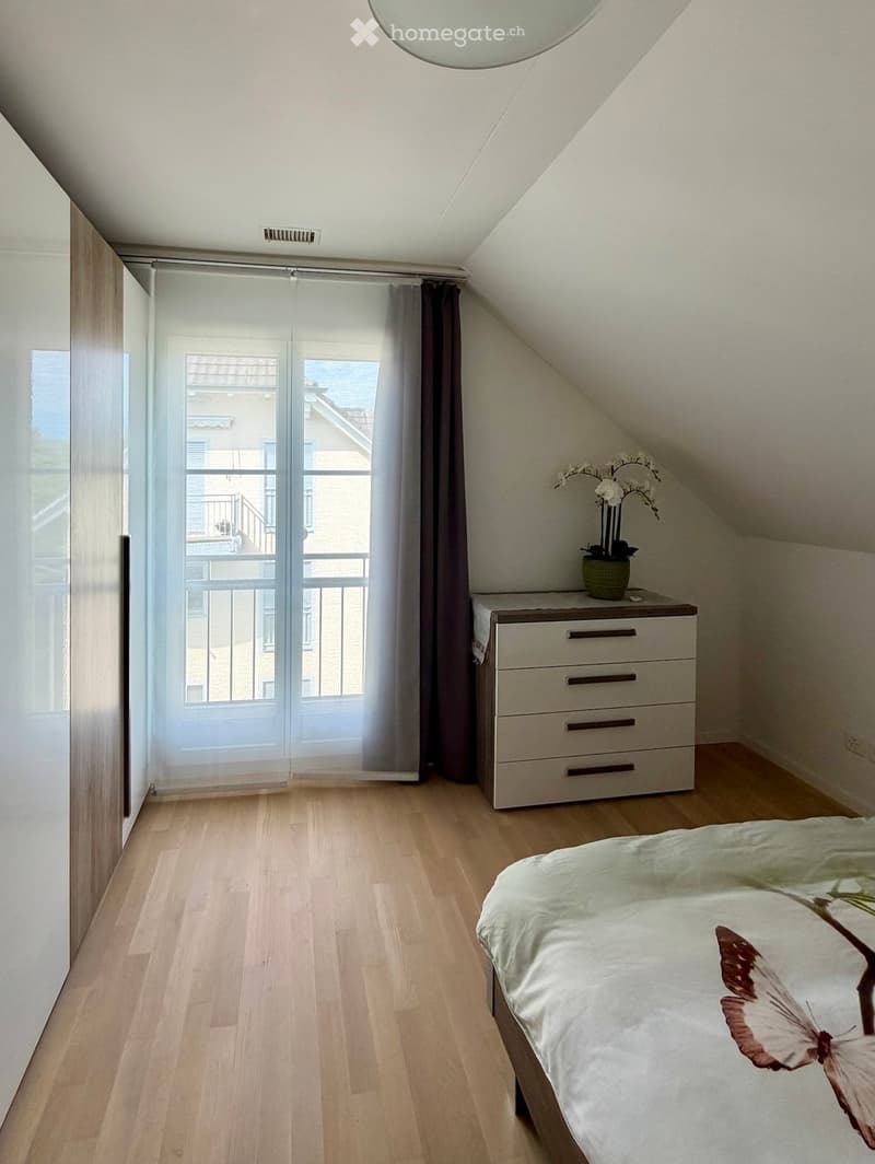 Bright bedroom with wooden flooring, large window offering a view, bed with decorative pillows, nightstand with a flower vase, and a wardrobe.