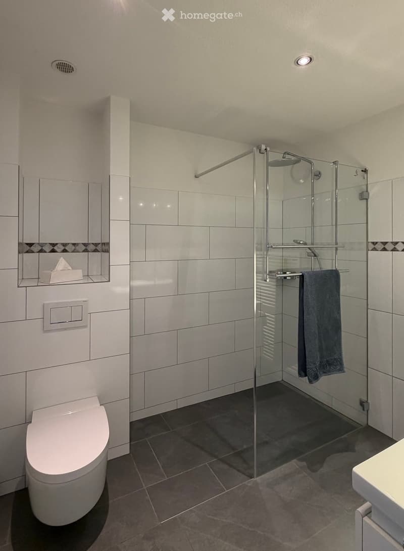 Modern bathroom with white tiles, a glass-enclosed shower, a toilet, a towel, and a mirror.