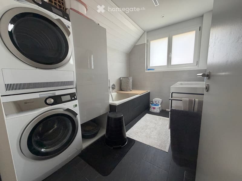 Modern bathroom with a washing machine, bathtub, sink, and a small stool. The space features grey tiles, a window, and a towel rack with towels.