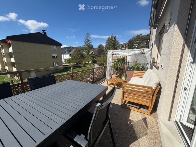 Balcony with a wooden sofa, a small table, and a dining table with chairs, overlooking a residential area with trees and buildings.