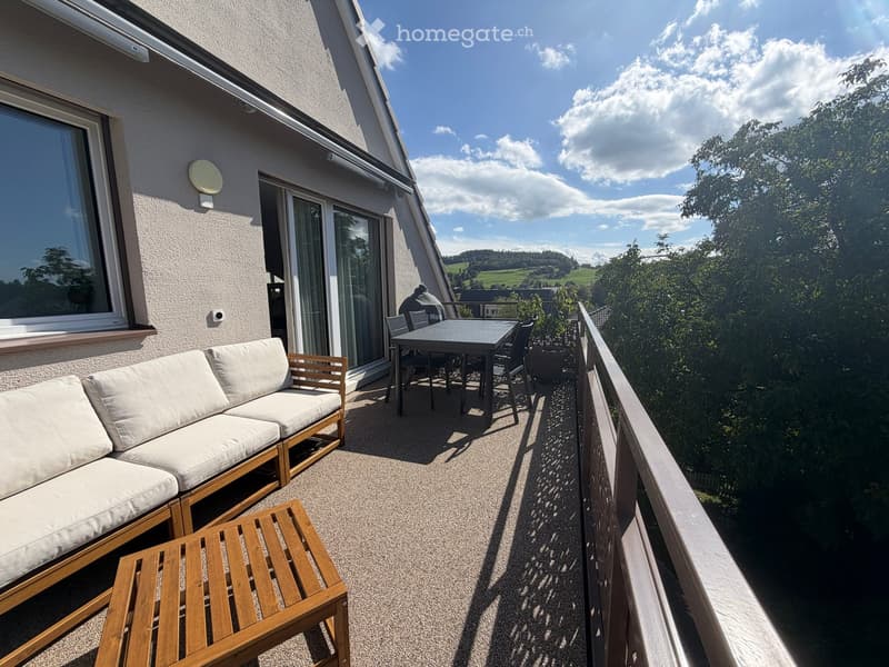 Balcony with a wooden sofa, small wooden table, and a dining table with chairs, overlooking a scenic landscape.