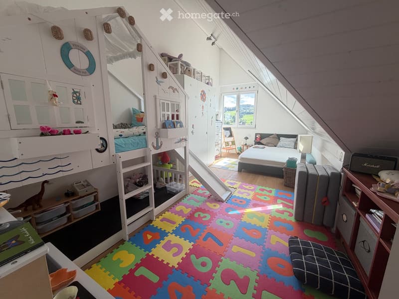 A well-lit, multi-functional bedroom with a bunk bed, a sofa, a colorful foam play mat, and various toys and storage items.