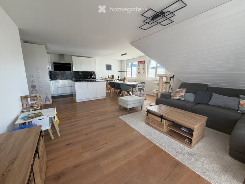 Open-plan living and dining area with wooden flooring, a grey sectional sofa, a wooden coffee table, an ottoman, a cat tree, a dining table with chairs, and a modern kitchen with white cabinets and a black countertop.